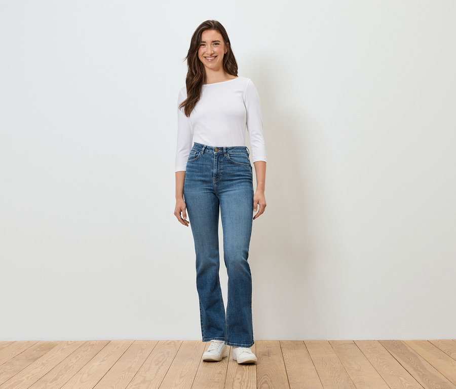 Eine Frau steht vor einer weißen Wand und trägt blaue Jeans, ein weißes Hemd und weiße Turnschuhe.