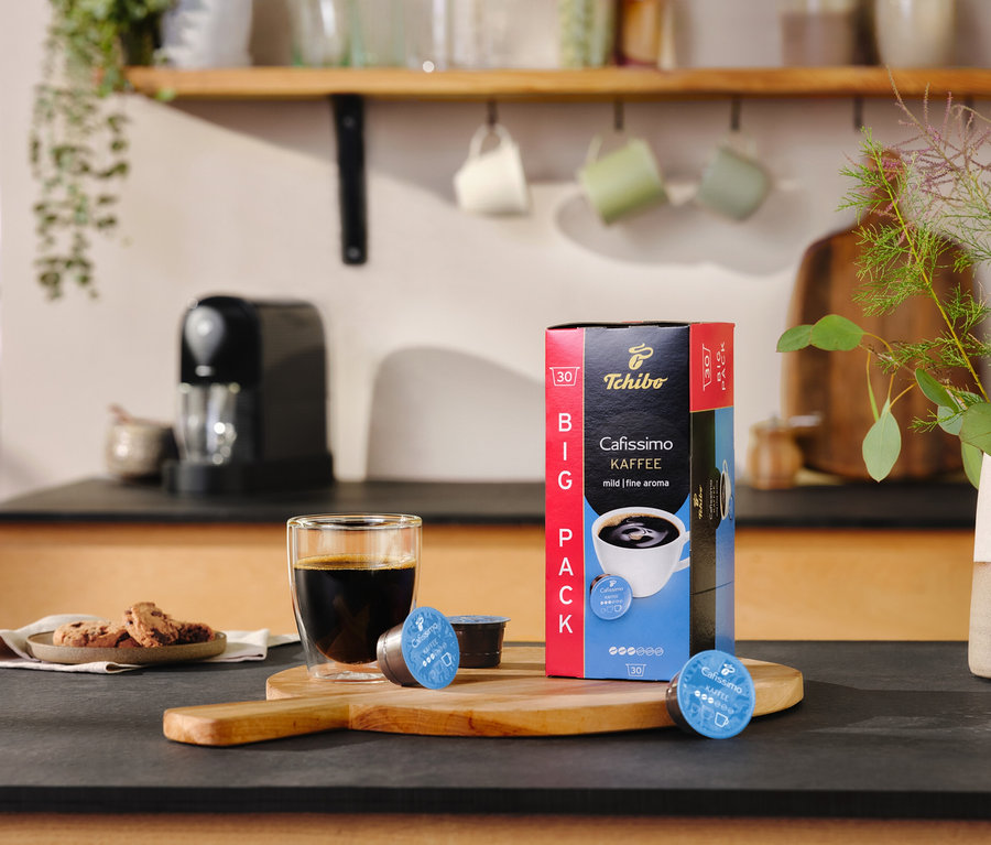 Auf einem Tisch stehen ein Glas Kaffee, eine Packung Cafissimo pure+, black und zwei Kapseln.