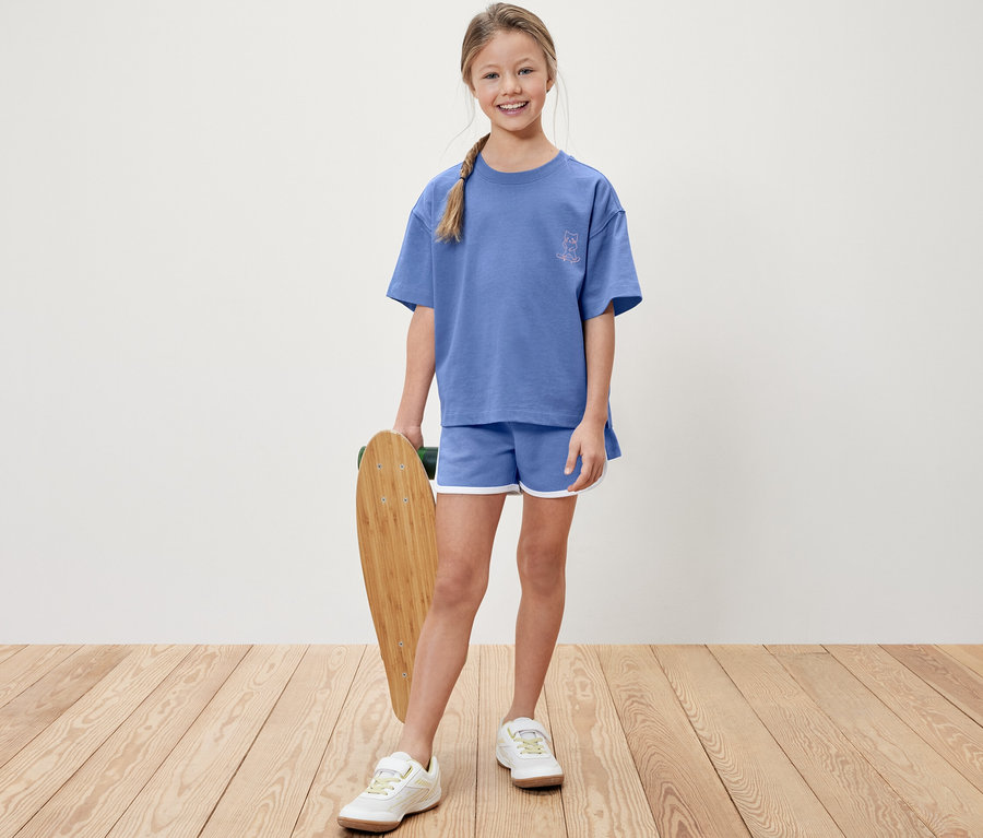 Mädchen hält ein Skateboard, trägt ein blaues T-Shirt und blaue Shorts.