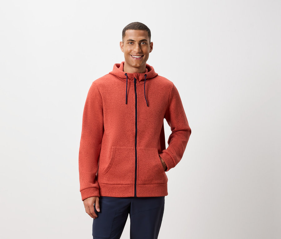 Ein Mann posiert vor einem weißen Hintergrund und trägt eine orangefarbene Kapuzenjacke mit Reißverschluss und dunkelblaue Hosen.