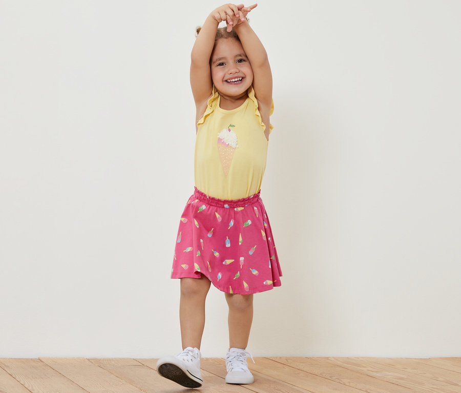 Lächelndes Mädchen steht mit erhobenen Händen und trägt ein gelbes T-Shirt und einen Kinder-Jerseyrock mit Hose, Eis.
