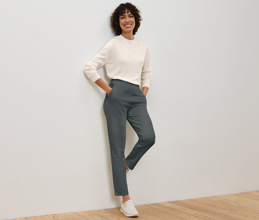 Eine Frau posiert an einer Wand, sie trägt eine graue Joggpants und einen Pullover.