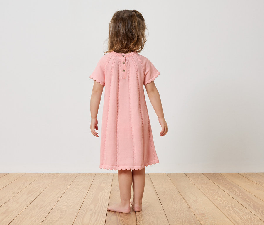 Ein Mädchen steht barfuß mit dem Rücken zur Kamera und trägt ein rosa Kleid.