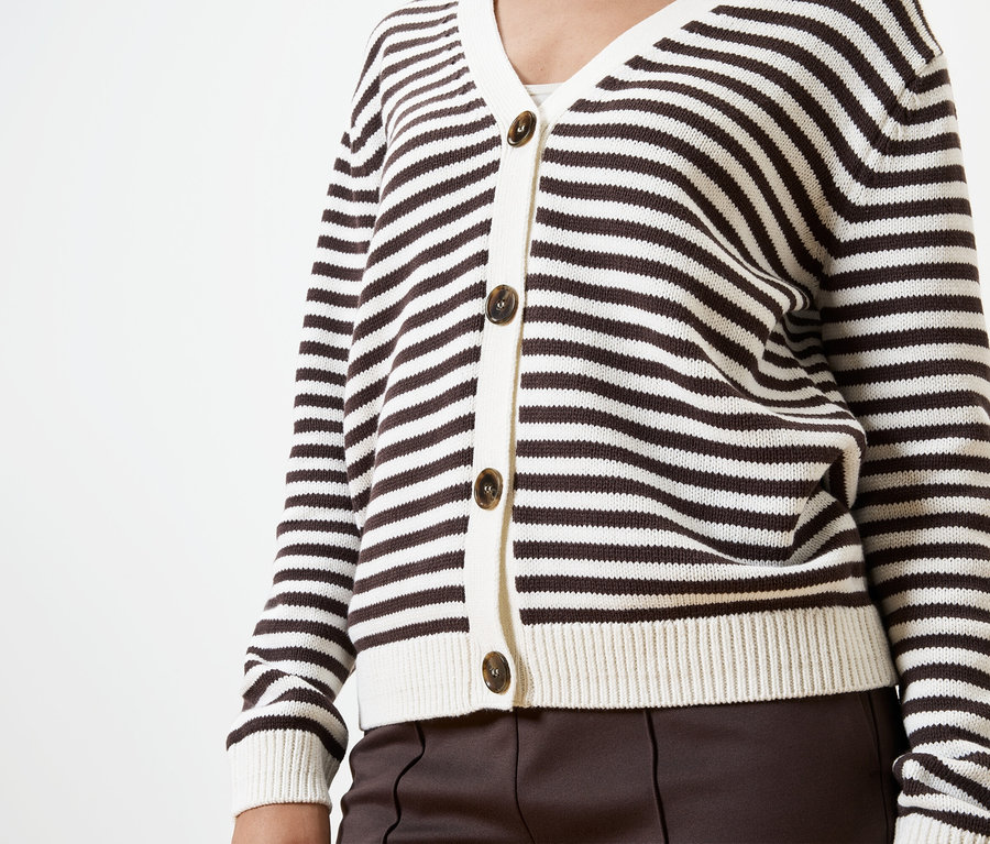 Oberkörper einer Person in einer braun-weiß gestreiften Strickjacke mit braunen Knöpfen und brauner Hose vor einem weißen Hintergrund.