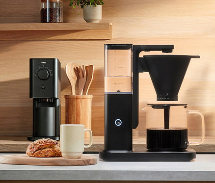 Eine Wilfa-Kaffeemühle und ein Kaffeebereiter stehen neben einer Tasse und einem Brotlaib auf einer Küchentheke.