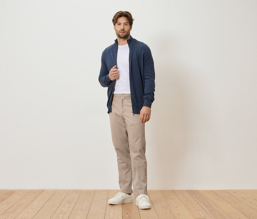 Ein Mann steht vor einer weißen Wand, er trägt eine marineblaue Strickjacke, ein weißes T-Shirt, beige Hosen und weiße Turnschuhe.