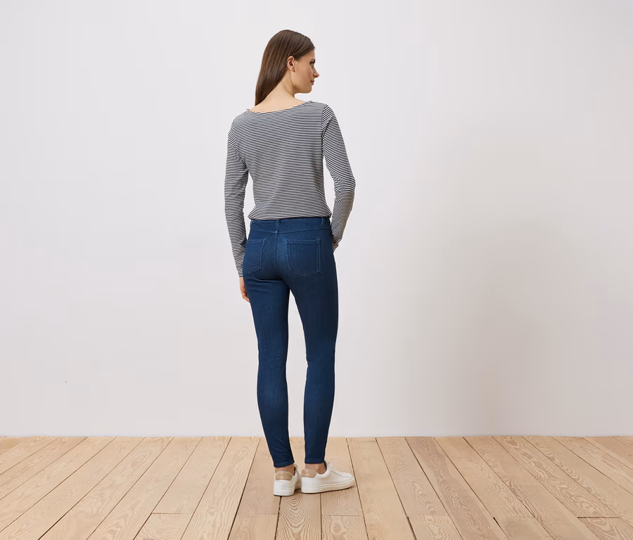 Eine Frau steht mit dem Rücken zum Betrachter, trägt Jeans, ein gestreiftes Langarmshirt und weiße Sneaker.
