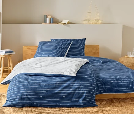 Bett mit blau-weiß gestreifter Bettwäsche und Knotenmuster. Auf dem Nachttisch stehen Bücher und eine Lampe in Schiffsform.