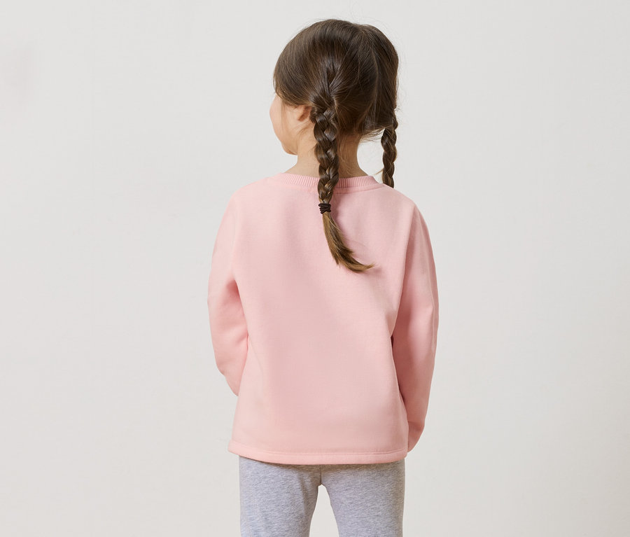 Rückansicht eines Mädchens mit Zöpfen, das einen rosa Pullover und graue Leggings trägt.
