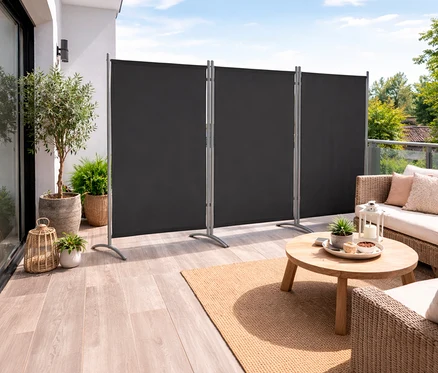 Auf einer Terrasse steht eine anthrazitfarbene, 3-teilige Leco Stellwand. Davor sind ein Tisch, ein Sofa und Pflanzen zu sehen.
