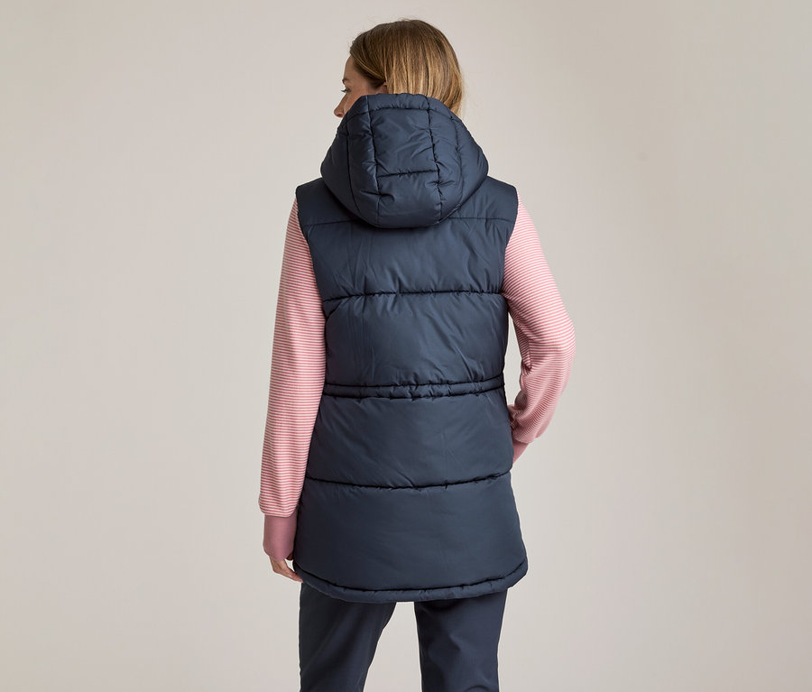Frau von hinten, die eine marineblaue, wattierte Weste mit Kapuze über einem rosa-weiß gestreiften Langarmshirt und dunklen Hosen trägt.