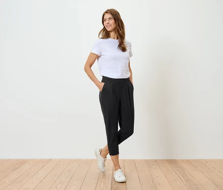 Frau mit weißem T-Shirt und schwarzer Hose posiert vor einer weißen Wand.