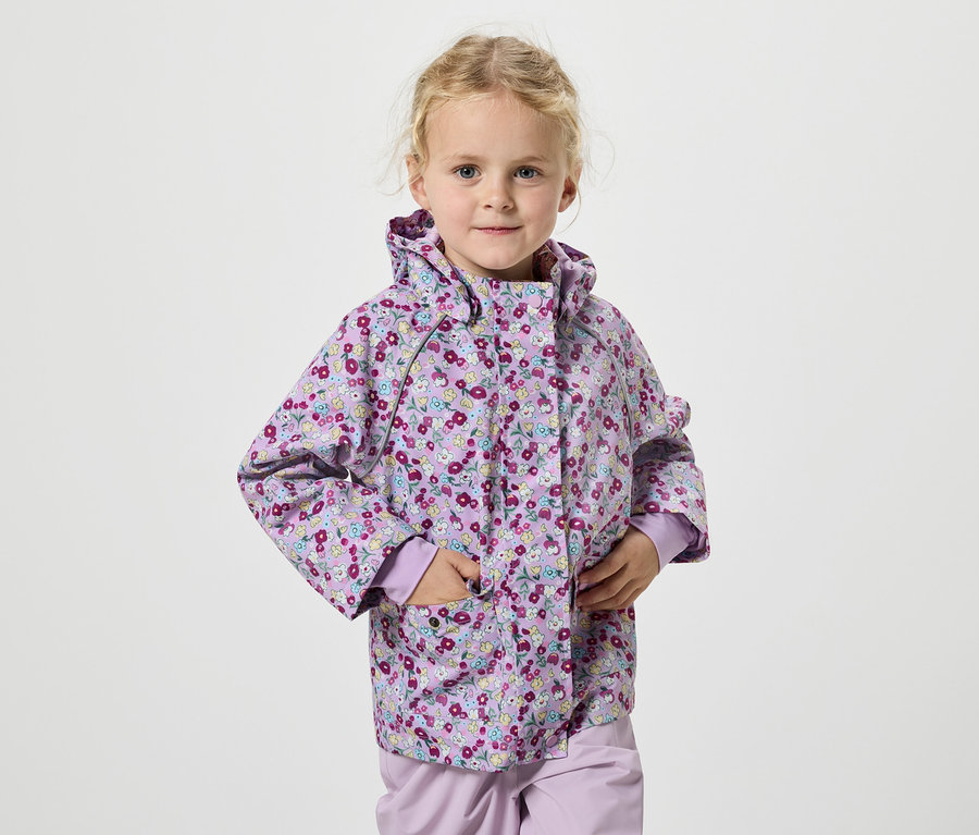 Mädchen mit blonden Haaren und lila Blumenjacke steht vor einem weißen Hintergrund.