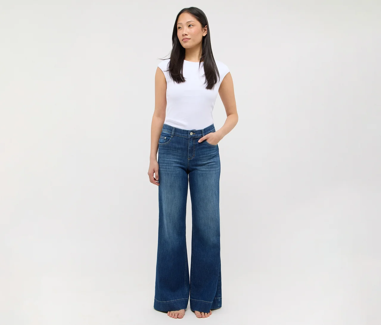 Eine Frau steht in einem weißen T-Shirt und einer blauen Jeans mit weitem Bein, die Hände in den Taschen.