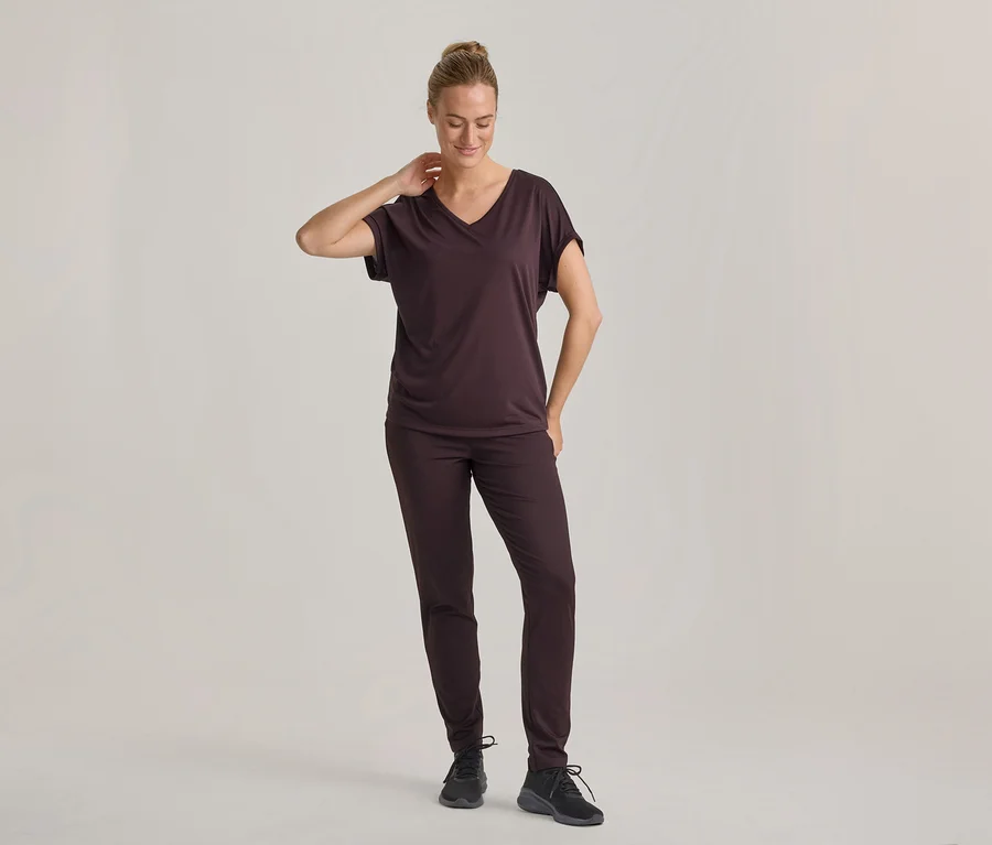 Frau in dunkelbraunem V-Ausschnitt-T-Shirt und passender Hose, mit schwarzen Turnschuhen, steht und blickt nach unten, eine Hand am Nacken.