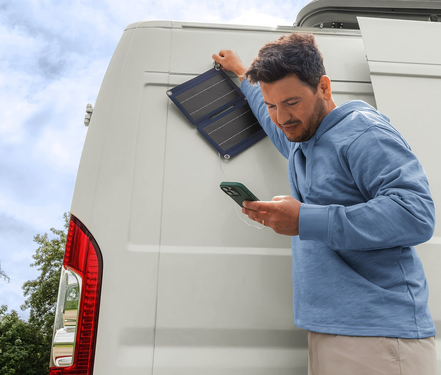 Mann in Sweat-Hoodie und beiger Hose lädt sein Handy mit einem faltbaren 10 W Solar-Ladegerät, das an einem weißen Camper befestigt ist.