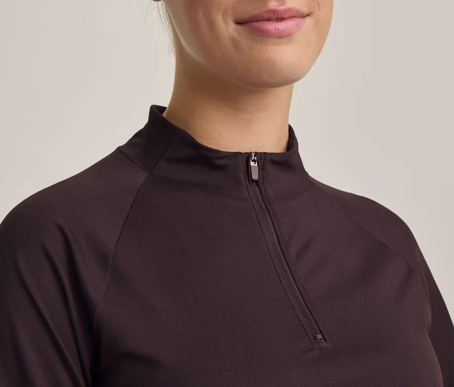 Nahaufnahme einer Person in einem dunkelbraunen Langarmshirt mit teilweise geöffnetem Reißverschlusskragen. Kinn und Mund der Person sind sichtbar, leicht lächelnd.