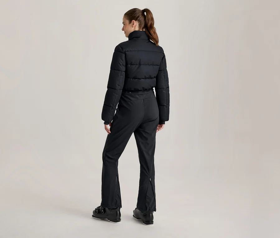 Frau von hinten, trägt eine schwarze Puffer-Skijacke, schwarze Skihose und schwarze Skischuhe. Ihre Haare sind zu einem Pferdeschwanz gebunden.