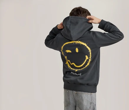 Eine Person von hinten, die einen dunkelgrauen Hoodie mit einem verwaschenen gelben Smiley-Gesicht auf dem Rücken und hellblaue Jeans trägt. Die Person hält die Kapuze mit beiden Händen.