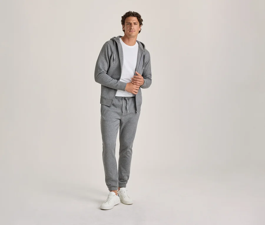 Ein Mann in einem grauen Kapuzenpullover, einem weißen T-Shirt und einer grauen Jogginghose steht mit weißen Turnschuhen und blickt in die Kamera.
