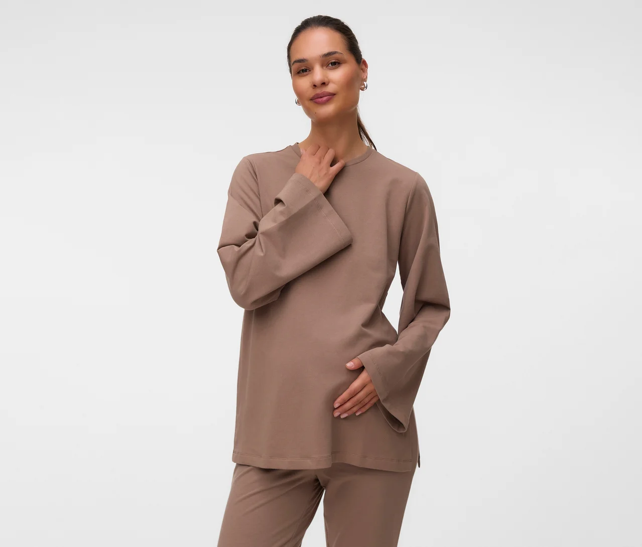 Eine schwangere Frau trägt ein hellbraunes MAMALICIOUS Sweat Top »MLSINE LS« und eine passende Hose, ihre rechte Hand liegt am Hals und die linke am Bauch.