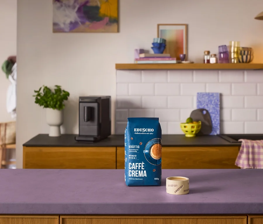 Blauer Beutel Eduscho Caffè Crema Kräftig Kaffeebohnen und eine kleine Eduscho Tasse auf lila Theke vor schwarzem »Esperto mini« Kaffeevollautomaten in einer Küche.