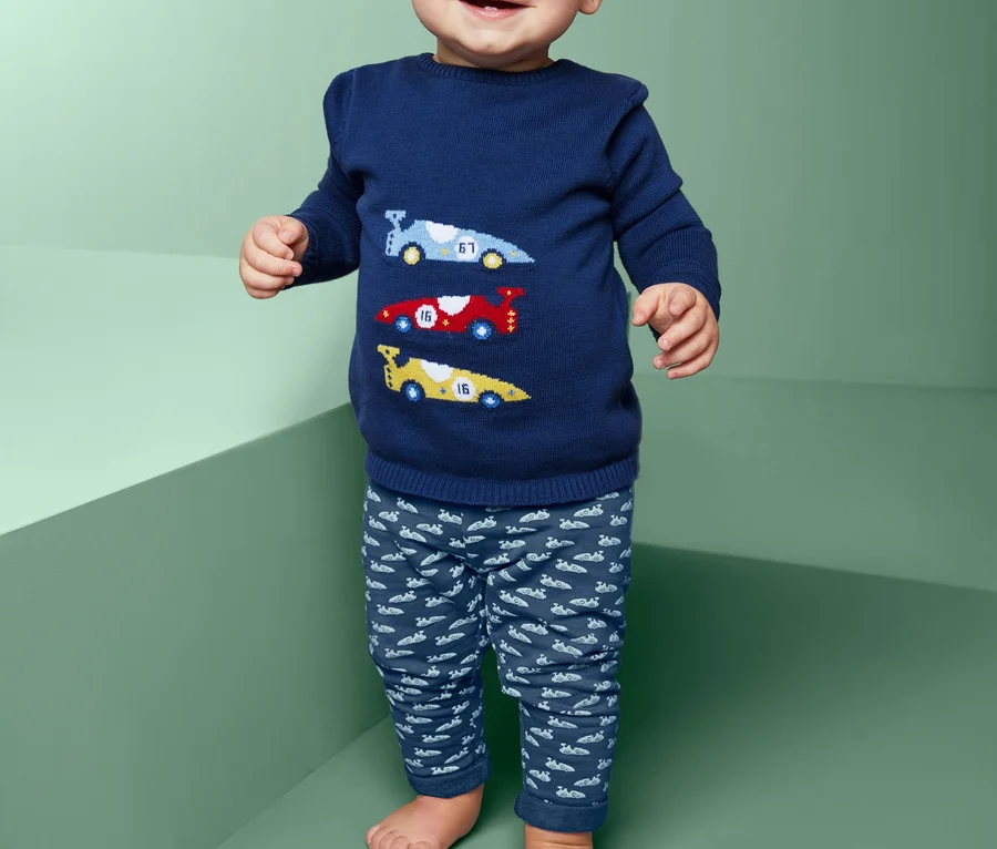 Lächelndes Baby steht und trägt einen blauen Pullover mit Autos und eine Sweathose.
