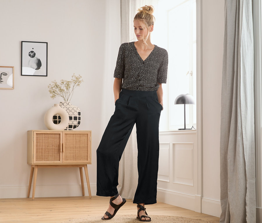 Frau steht im Raum und trägt eine Satin-Bundfaltenhose und ein gemustertes Oberteil.
