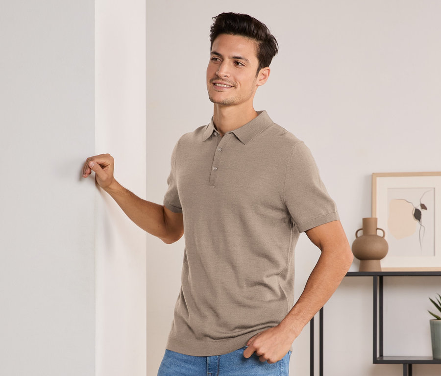 Mann lehnt an Wand, trägt beige meliertes Merino-Poloshirt, Jeans und weiße Sneaker.