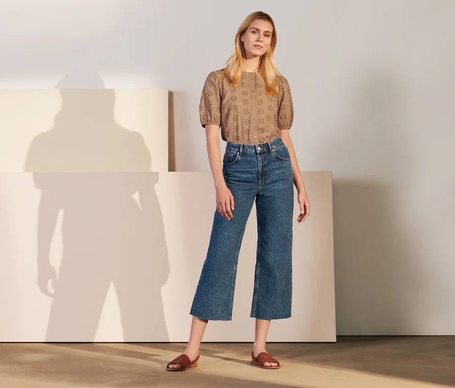 Eine Frau posiert stehend in Culotte-Jeans und brauner Bluse.