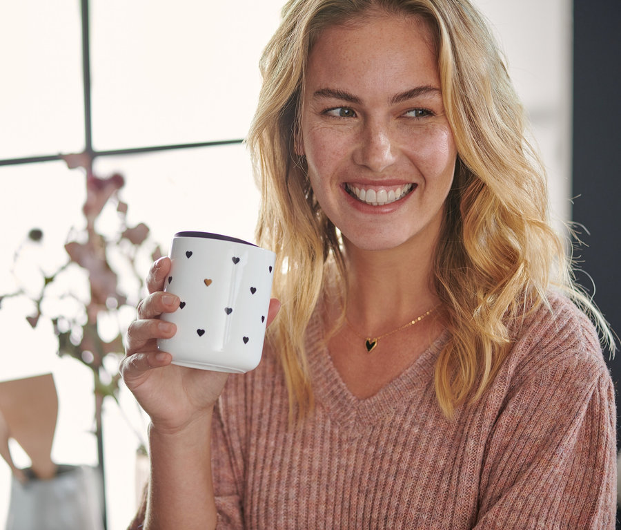 Frau hält einen Porzellanbecher mit Deckel und Herzchenmuster.
