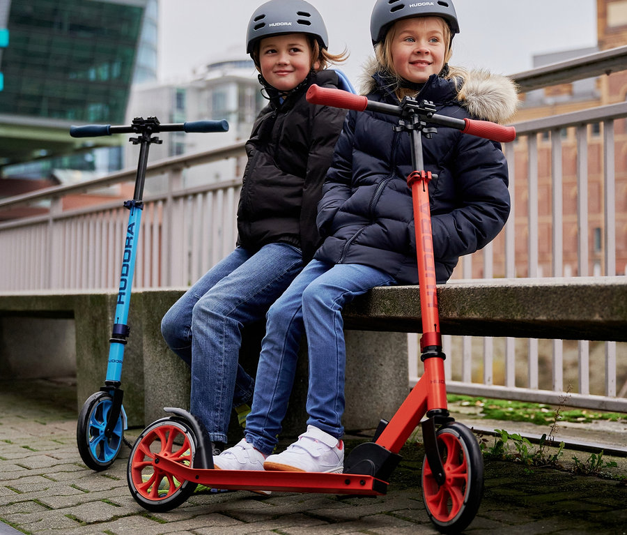 Zwei Kinder sitzen draußen nebeneinander auf einer Bank, neben ihnen stehen HUDORA »Up 200« Scooter in Rot und Blau.