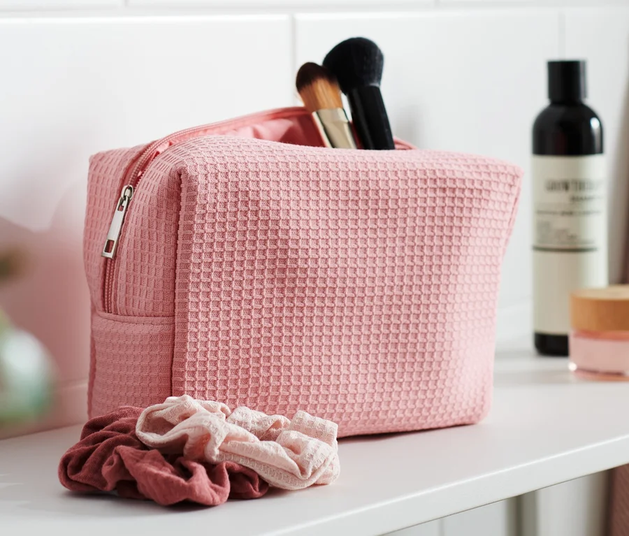 Eine rosa Kulturtasche mit Kosmetikpinseln steht auf einem weißen Regal. Daneben liegen zwei Haargummis.