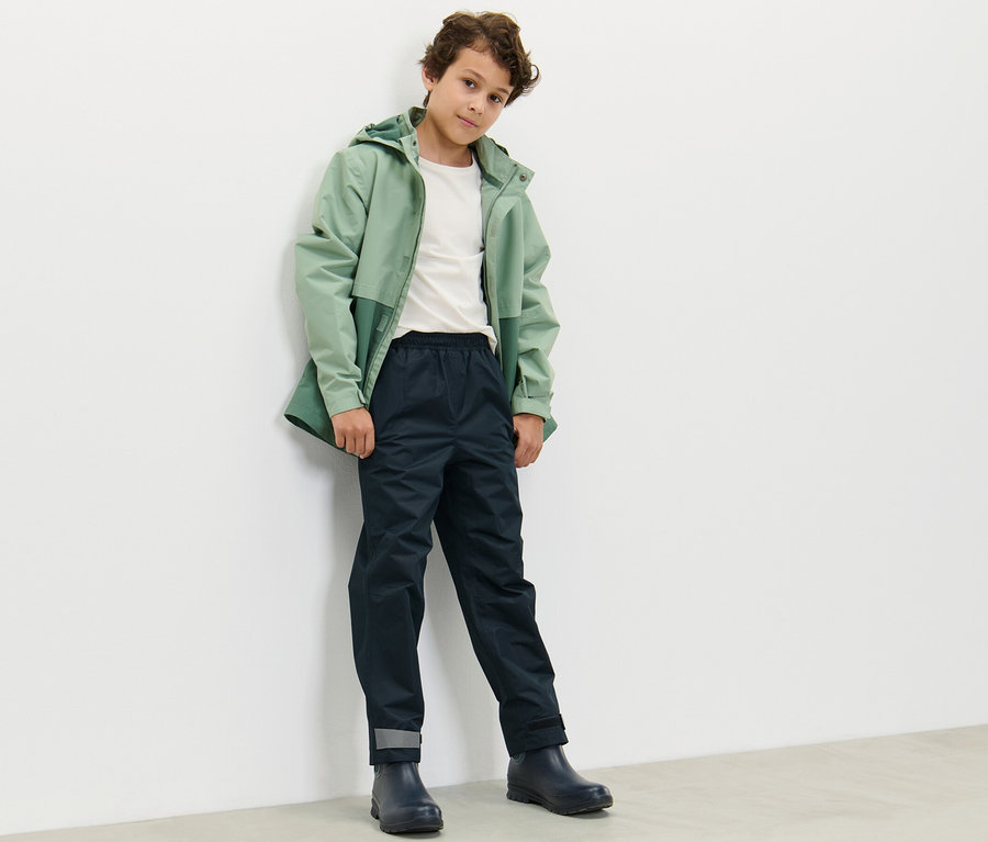Ein Junge in einer grünen Jacke und dunkelblauer Kinder-Regenhose steht an einer weißen Wand.