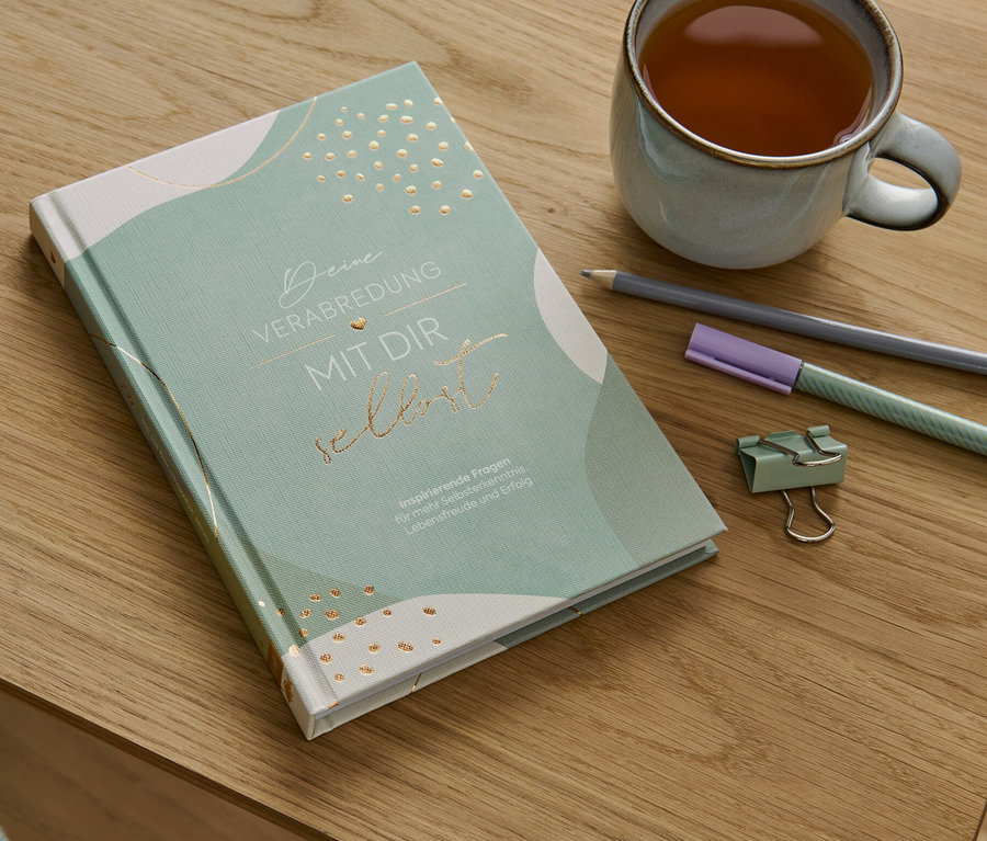 Auf einem Holztisch liegen das Buch »Deine Verabredung mit dir selbst«, eine Tasse Tee, ein Bleistift, ein Textmarker und eine Büroklammer.