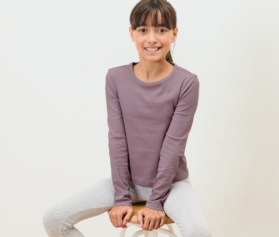 Mädchen sitzt auf einem Hocker und trägt ein Kinder-Langarmshirt mit Rippstruktur.