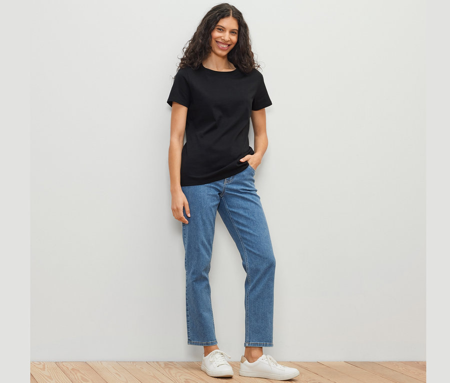 Frau mit schwarzem Basic T-Shirt, blauen Jeans und weißen Schuhen steht vor einer weißen Wand.