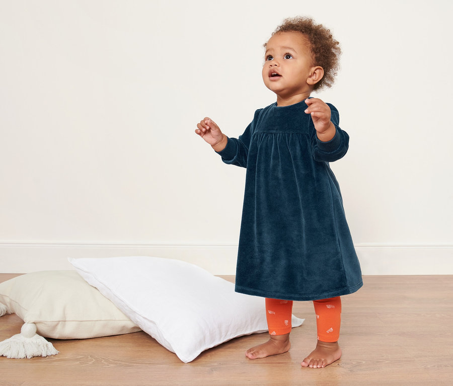 Baby steht auf dem Boden neben Kissen und trägt ein Baby-Nickikleid.