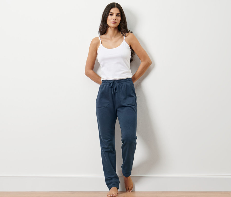 Frau steht an einer Wand, trägt ein weißes Top und eine Loungewear-Hose in Navy.