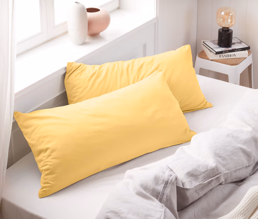 Zwei gelbe Jersey-Kopfkissenbezüge liegen auf einem Bett mit weißen Laken.