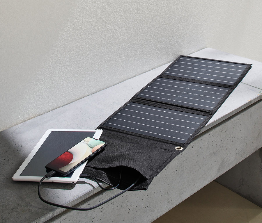 Ein Solar-Ladegerät, faltbar, lädt ein Handy, das auf einem Tablet auf einer Betonbank liegt. Daneben befindet sich ein Rucksack.