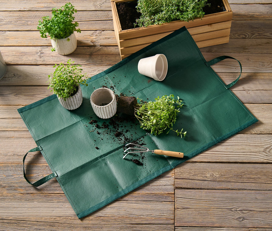 Eine Gartenabfalltasche liegt auf einem Holzboden neben Topfpflanzen und einer Gießkanne.