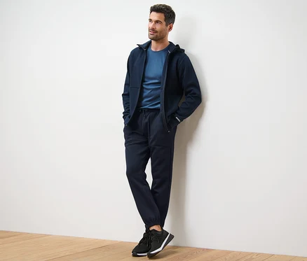 Mann lehnt an Wand, trägt dunkelblaue Kapuzenjacke, T-Shirt und Funktions-Sweathose.
