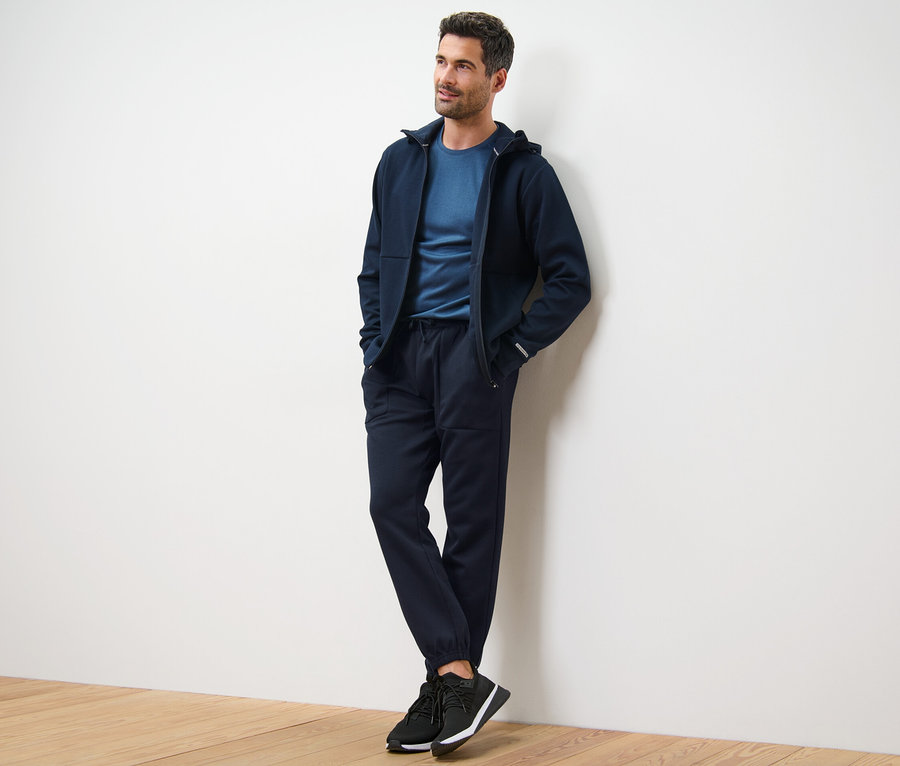 Mann lehnt an Wand, trägt dunkelblaue Kapuzenjacke, T-Shirt und Funktions-Sweathose.