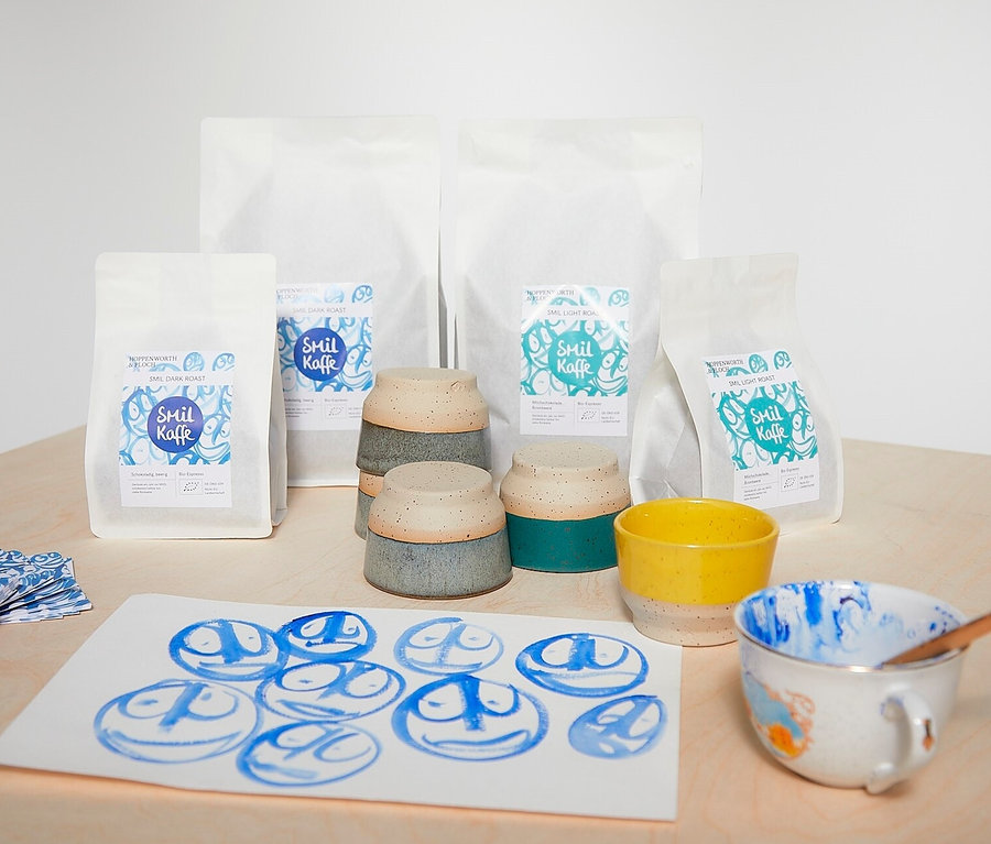 Auf einem Tisch stehen vier Packungen Hoppenworth & Ploch - Smil - Dark Roast Omni-Roast, Keramiktassen und Malereien mit blauen Gesichtern.