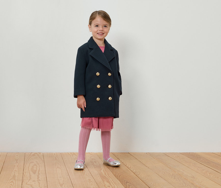 Mädchen steht in marineblauem zweireihigem Mantel, rosa Kleid und Strumpfhose.