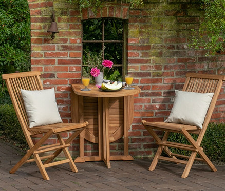 Zwei Holzstühle mit weißen Kissen stehen an einem deVries Balkontisch, Teak, 60 x 120 auf einer gepflasterten Terrasse.