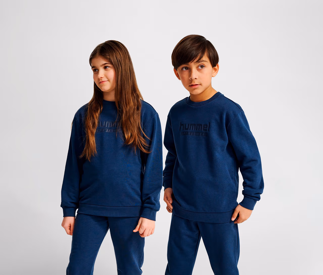 Zwei Kinder stehen nebeneinander und tragen blaue Hummel HMLJR Base-Sweatshirts und blaue Jogginghosen.