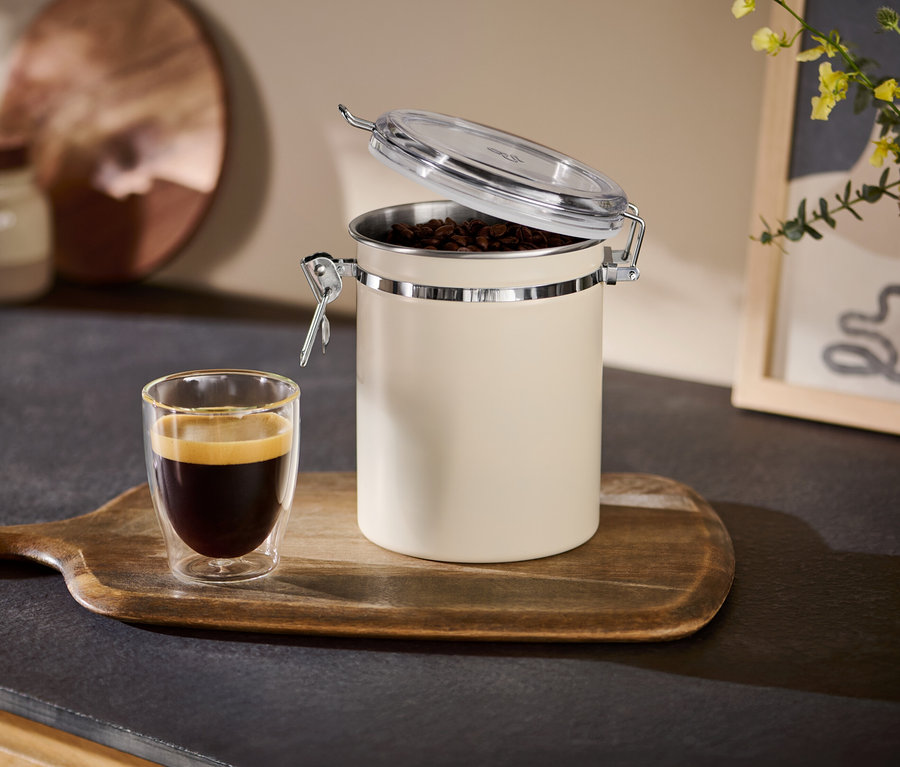 Geöffnete beige Aromadose neben einer Tasse Espresso auf einem Holztablett.