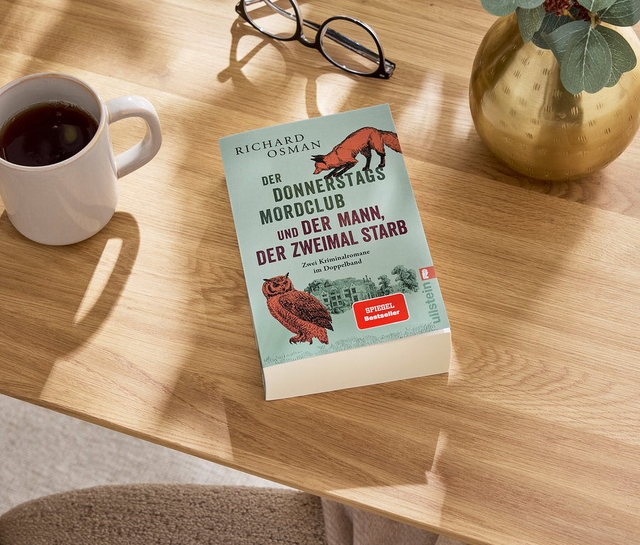 Auf einem Tisch liegen ein Buch, eine Tasse mit einem Getränk, eine Brille und eine Vase.